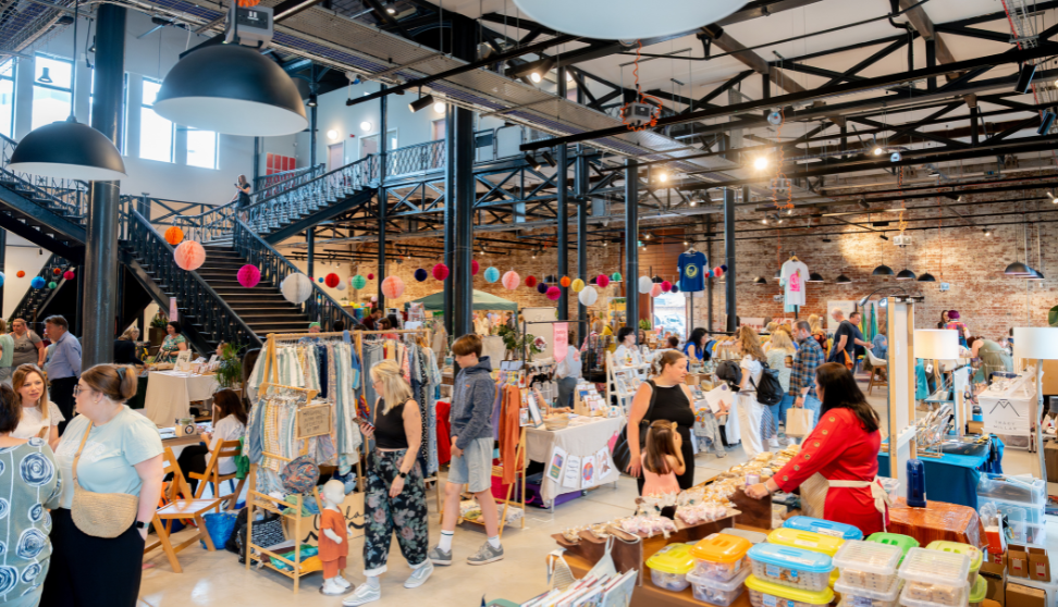 Indoor craft and makers market in a large industrial hall, with stalls of handmade goods and food, shoppers browsing, and colorful decorations hanging by a metal staircase.